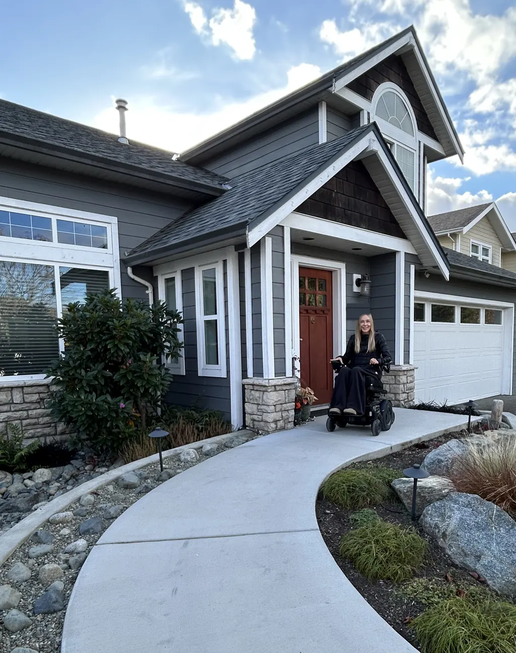 A woman in a power wheelchair smiles at her front door, at the end of a wide, level concrete path through the garden.